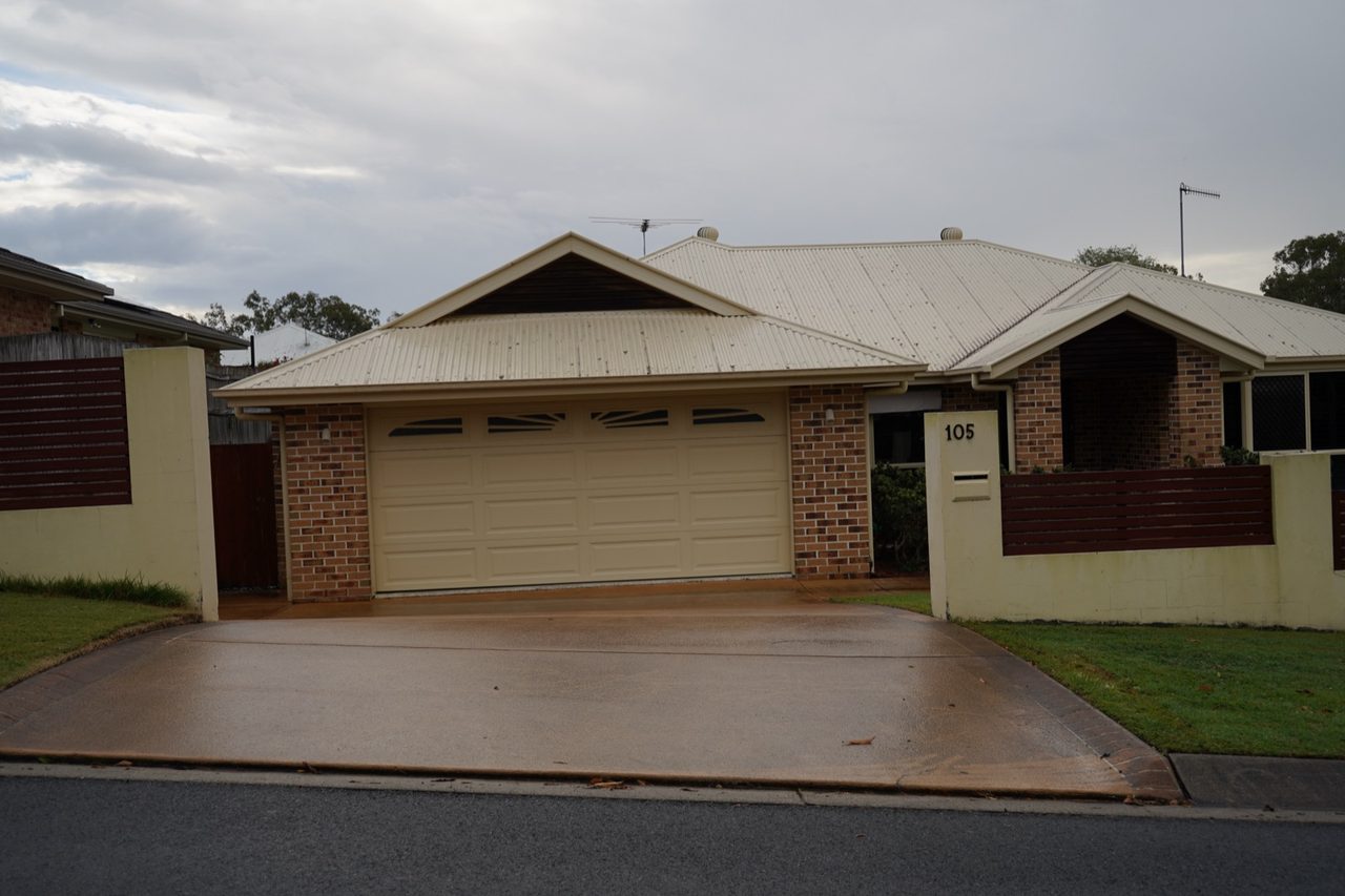 Plain concrete driveway with patchy tan and brown staining in front of a brick home's cream double garage in Gumdale, Brisbane