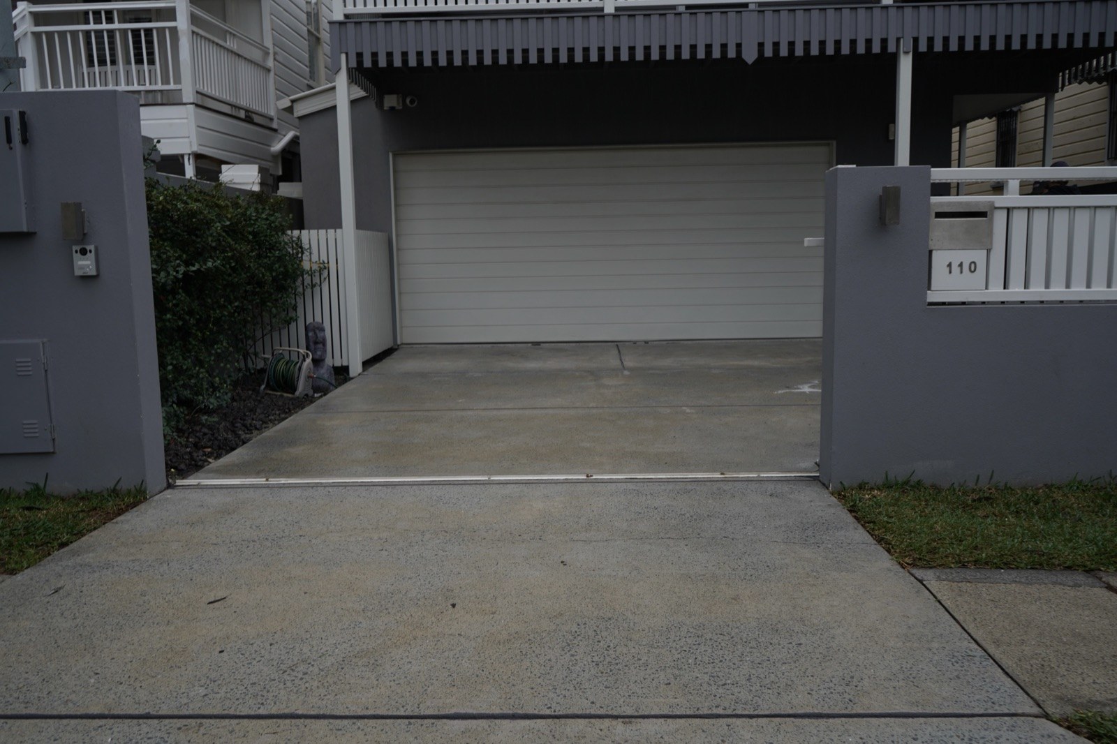 Worn pale grey concrete driveway before resurfacing, running between rendered gate piers to a white garage door beneath a Queenslander-style home's deck